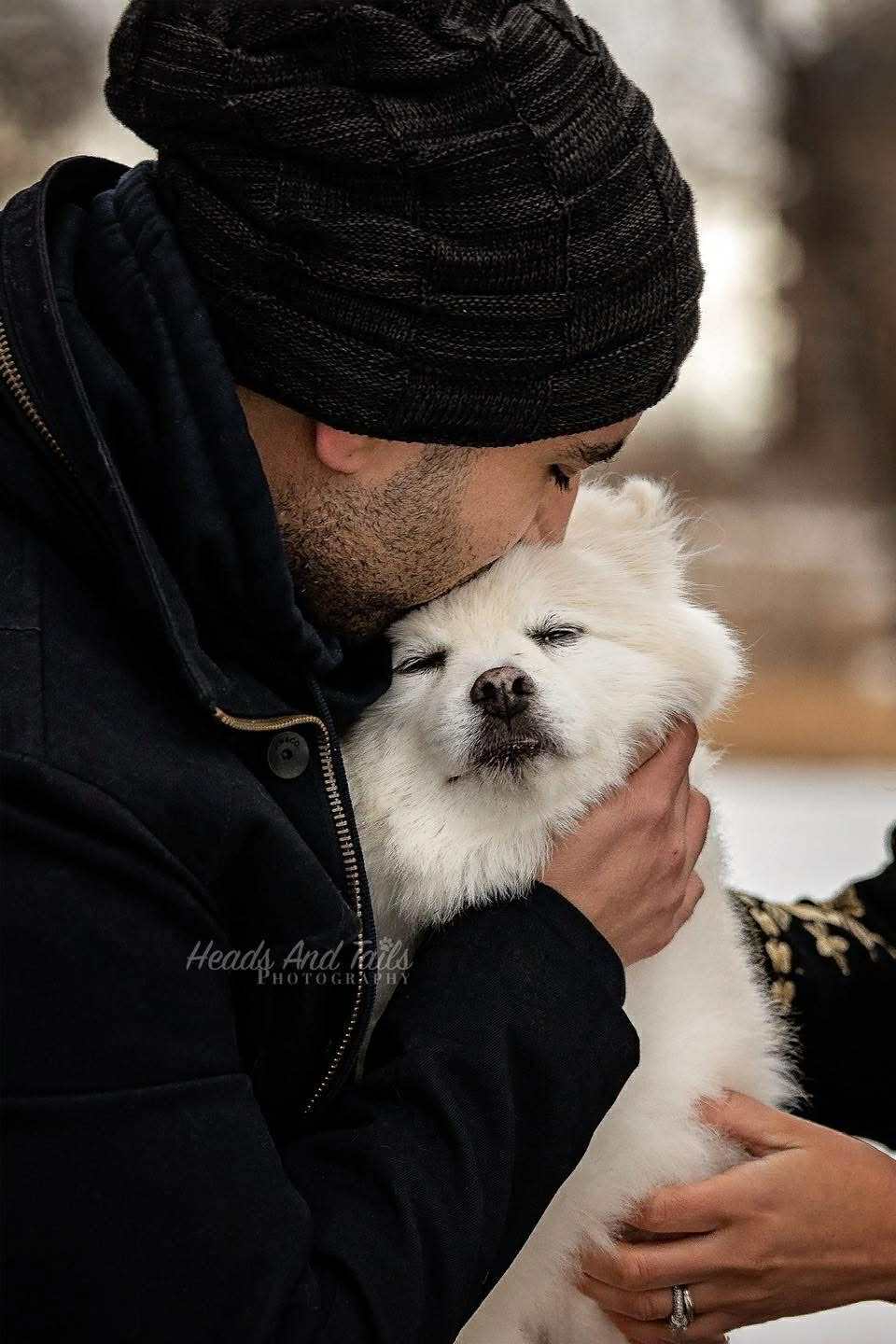 Winter Cuddle – Senior Dog & Owner | Nova Scotia Pet Photography Tender winter portrait of a man in a knit beanie gently kissing his senior white fluffy dog, captured by Nova Scotia pet photographer Heads and Tails Photography.